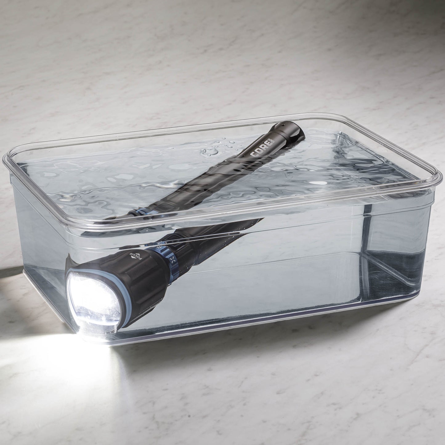 Dark gray CORE 5000 lumen rechargeable flashlights with blue accents turned on in a clear container filled with water.