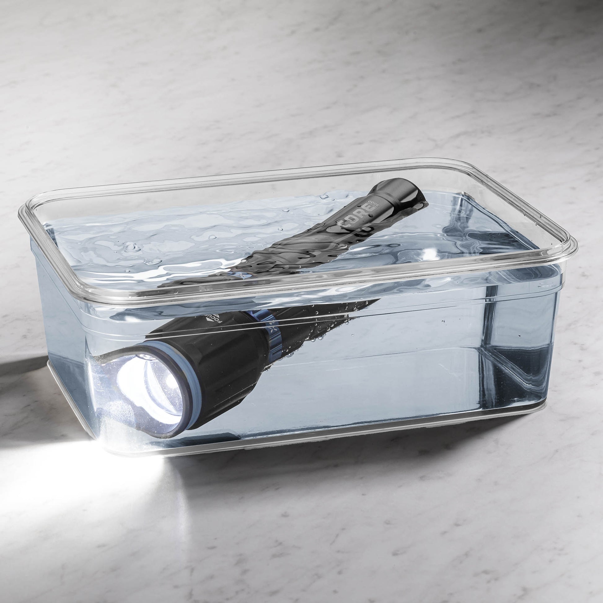 Dark gray CORE 3000 lumen rechargeable flashlights with blue accents turned on in a clear container filled with water. 