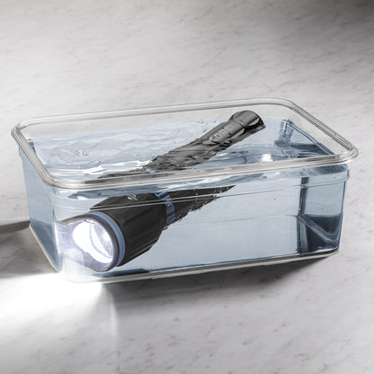 Dark gray CORE 3000 lumen rechargeable flashlights with blue accents turned on in a clear container filled with water. 