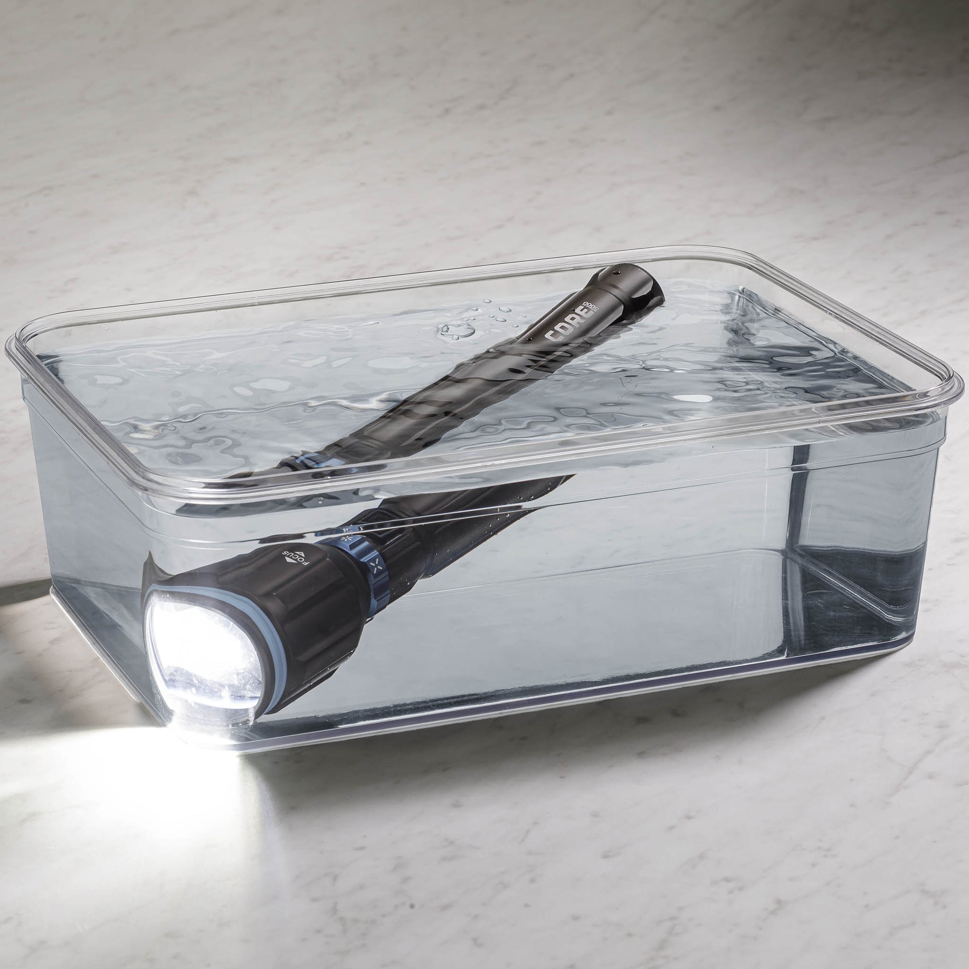 Dark gray CORE 5000 lumen rechargeable flashlights with blue accents turned on in a clear container filled with water. 